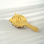A light natural bamboo hairbrush with small brown bristles, featuring an engraved logo and text on the back of the head, resting on a white surface with soft diagonal shadows.