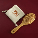 A natural bamboo hairbrush with soft bristles, shown next to a beige drawstring pouch with the brand logo 'OU' on a dark burgundy background.