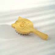 A light natural bamboo hairbrush with small brown bristles, featuring an engraved logo and text on the back of the head, resting on a white surface with soft diagonal shadows.