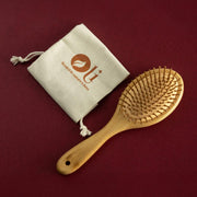 A natural bamboo hairbrush with soft bristles, shown next to a beige drawstring pouch with the brand logo 'OU' on a dark burgundy background.
