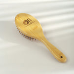 A light natural bamboo hairbrush with a wooden handle and metal bristles, featuring an engraved logo and text on the head, set against a softly lit white background.