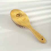 A light natural bamboo hairbrush with a wooden handle and metal bristles, featuring an engraved logo and text on the head, set against a softly lit white background.