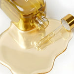 A close-up of a clear glass bottle filled with golden yellow liquid, tipped over so the liquid pools in a wavy shape beneath it. A glass dropper with a gold cap rests on the surface, and the bottle cap is partially visible.